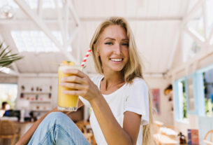romantic woman in blue jeans drinking orange cocktail with pleasure indoor shot of smiling blonde girl holding glass of cold juice while sitting in cafeteria