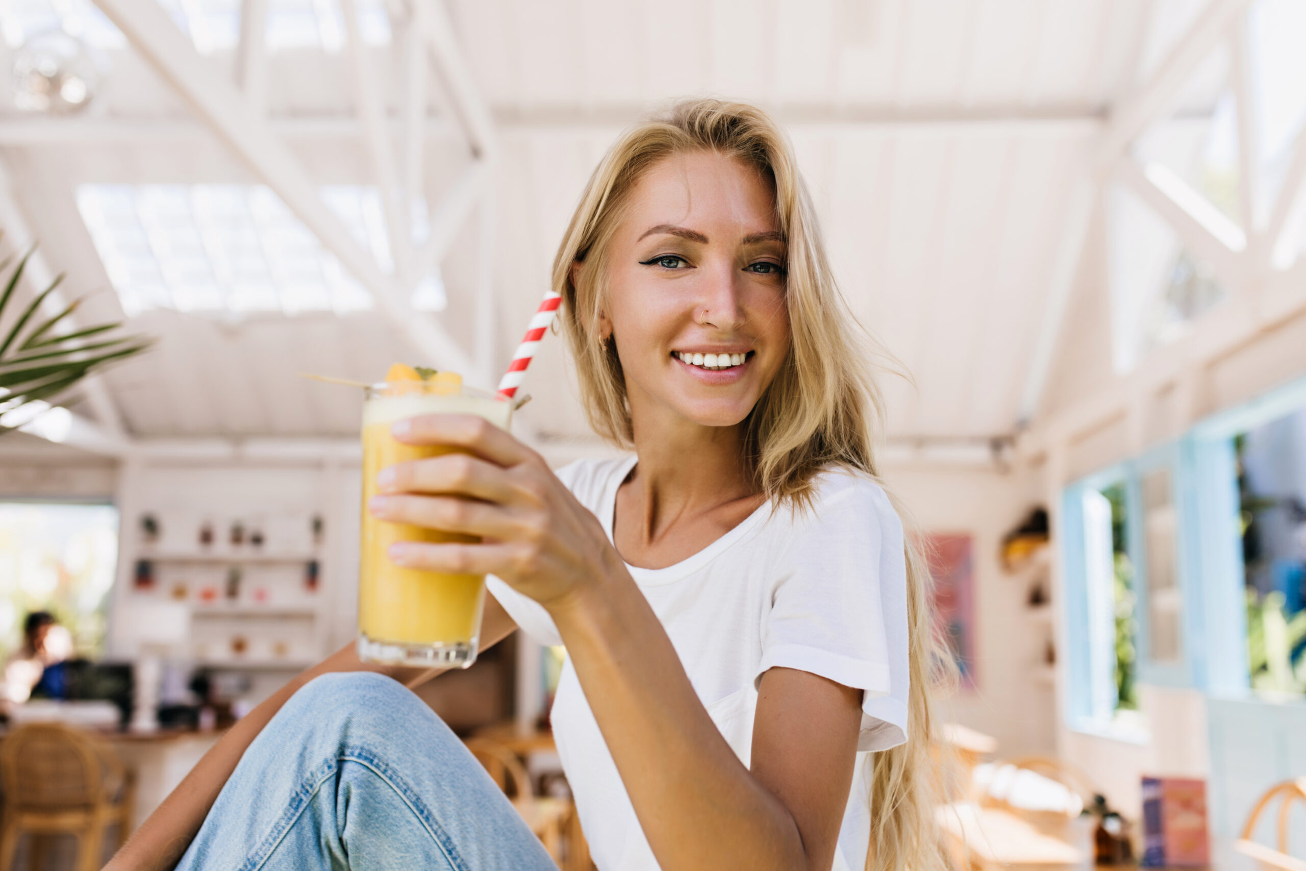 romantic woman in blue jeans drinking orange cocktail with pleasure indoor shot of smiling blonde girl holding glass of cold juice while sitting in cafeteria scaled